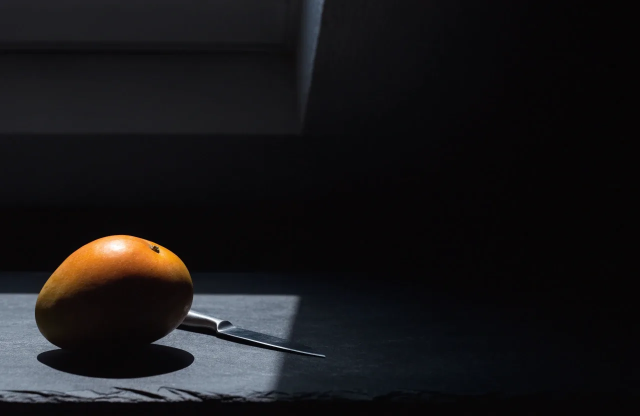 Prepola mango na mat crnoj plohi uz mali nož, desna strana kadra u dubokoj sjeni